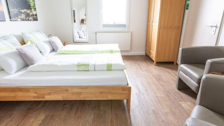 A bright room with two beds and wooden flooring. There is a chair and a wardrobe, as well as a mirror and a window.