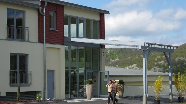 A modern building with large windows and a covered entrance area. In the foreground, some plants and two people can be seen talking.