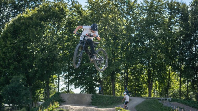 A BMX rider jumps in the air over a jump ramp. In the background, green trees are visible.
