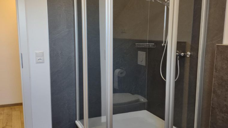 Een moderne douchecabine met heldere glazen wanden staat in een lichte badkamer. De vloer is met houten planken gelegd, en er is een deur die naar een andere kamer leidt.