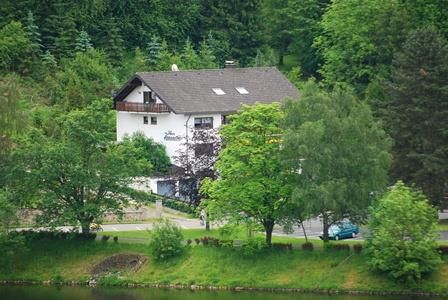 Een rustig woonhuis omringd door groene bomen. Op de voorgrond loopt een smal pad langs een water.