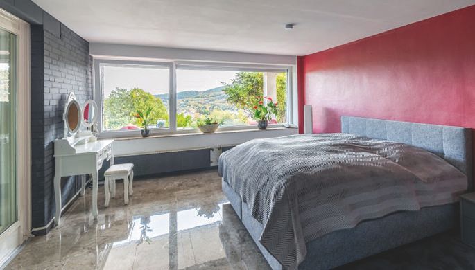 A modern bedroom with a large bed and windows overlooking the landscape. The walls are painted in soft colors, and there is an elegant vanity table.