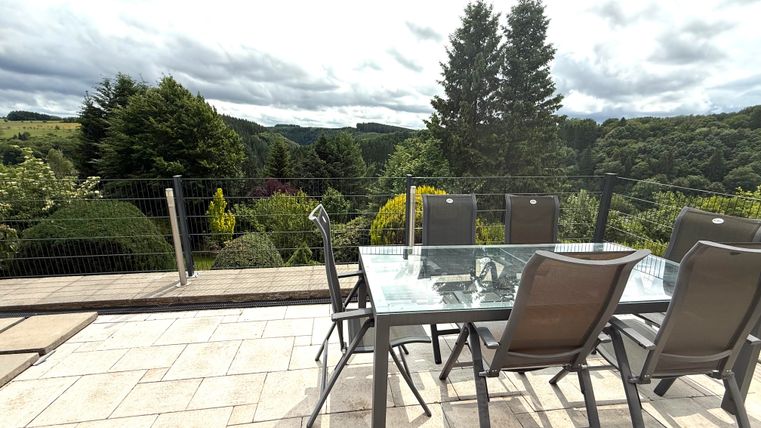 Een moderne eetruimte buiten met een tafel en stoelen op een terras. Op de achtergrond zijn groene heuvels en bomen te zien, onder een bewolkte lucht.