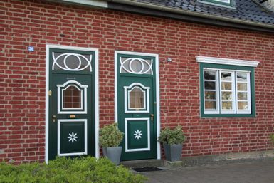 Eine charmante rote Backsteinwand mit grünen Türen und Fenstern. Vor den Türen stehen Töpfe mit Pflanzen.