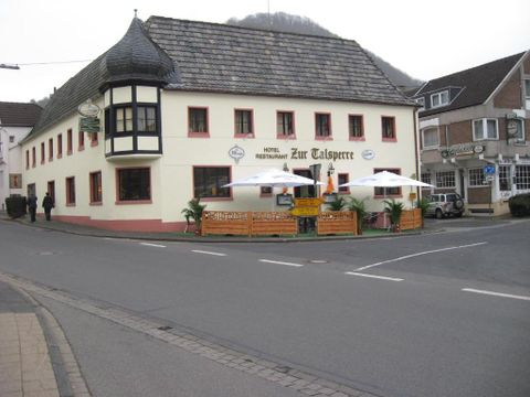 A charming restaurant and hotel named "Zur Talsperre" at a street corner. It has an inviting terrace with sun umbrellas and is surrounded by a quiet street.