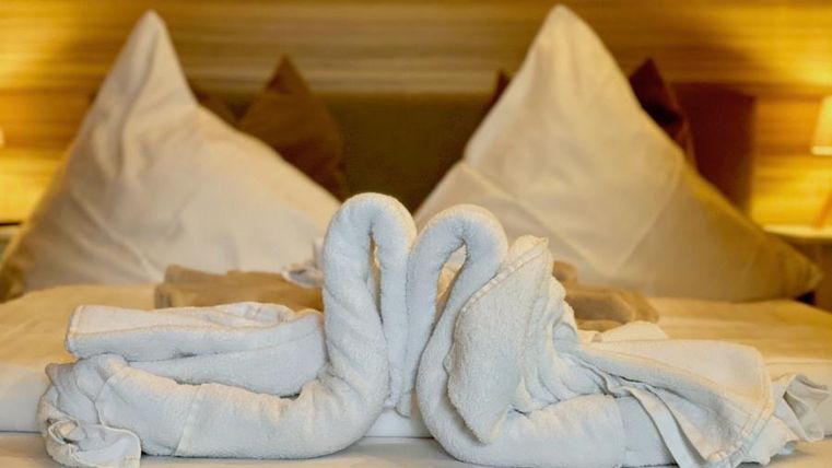 Ein schön dekoriertes Bett mit zwei handtuchgefalteten Schwänen in der Mitte. Im Hintergrund sind Kissen und sanftes Licht zu sehen.