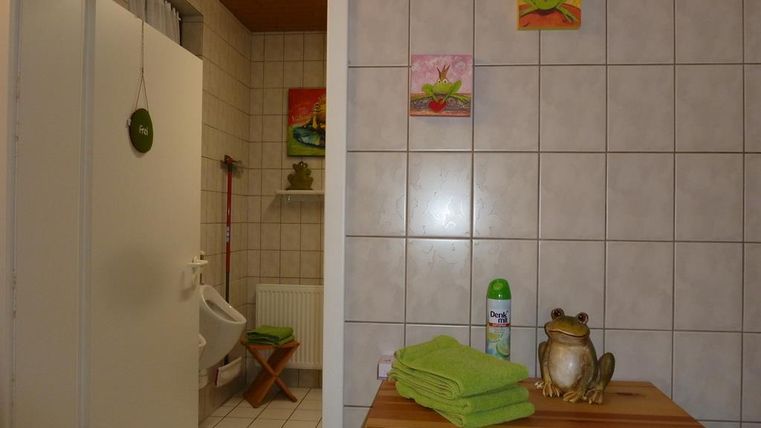 A bathroom with white tiles and a bright area. There are green towels, a fruit bottle, and decorative pictures on the wall.