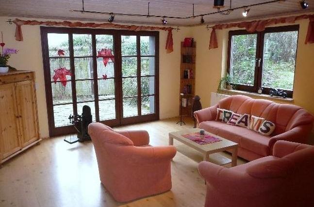 Ein gemütliches Wohnzimmer mit rosa Sesseln und einem Tisch in der Mitte. Große Fenster bieten einen Blick auf den Garten.