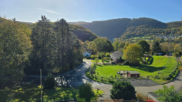 A picturesque landscape with green meadows and trees. In the background, gentle hills and a blue sky are visible.