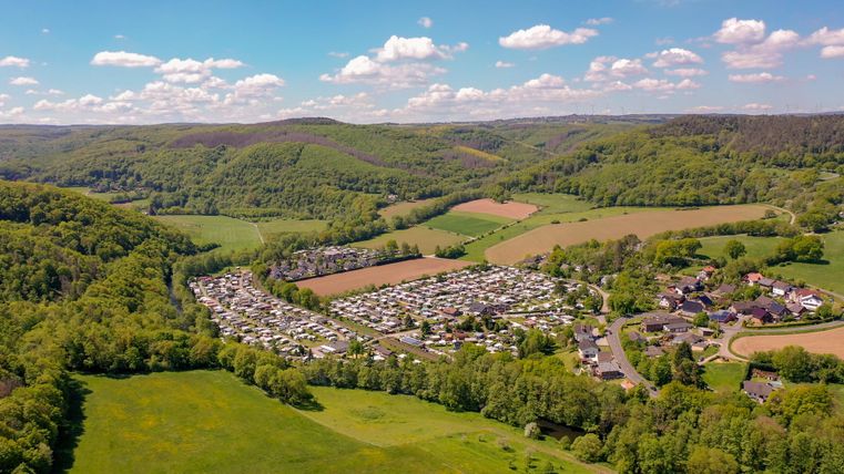 Eine Luftaufnahme eines Campingplatzes umgeben von grünen Hügeln und Wiesen. Der Himmel ist klar mit einigen Wolken.