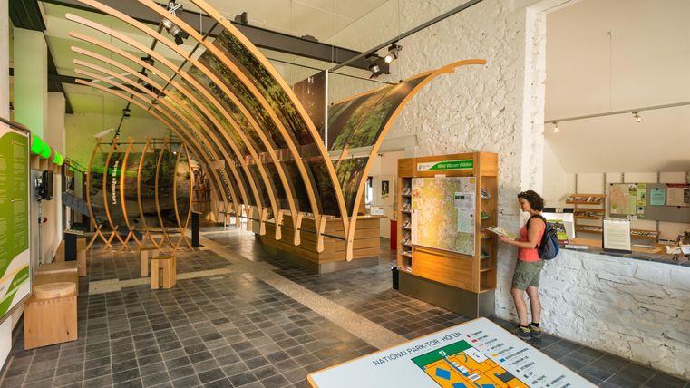 Een moderne ingang van een informatiecentrum met houten structuren. Op de voorgrond zijn bezoekers te zien bij de informatiebalie.