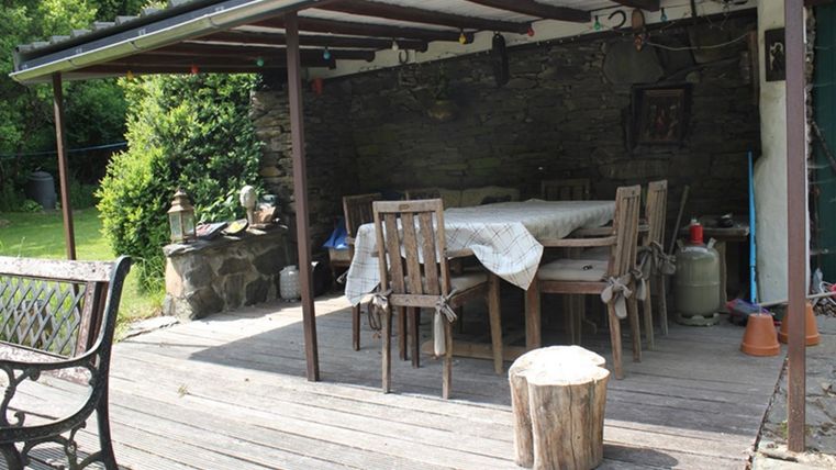 A cozy covered outdoor dining area with a table and chairs. Surrounded by green plants and a wooden log as a stool.
