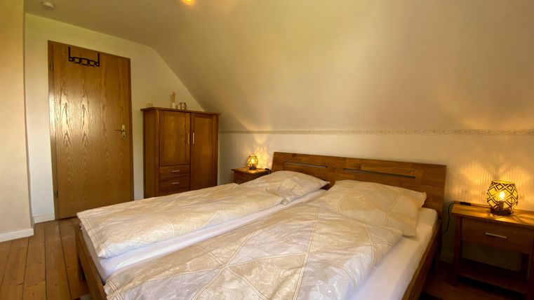 Een gezellige slaapkamer met een tweepersoonsbed, houten vloer en warm licht. Aan de muur staat een houten kast en een nachttafeltje met een lamp.