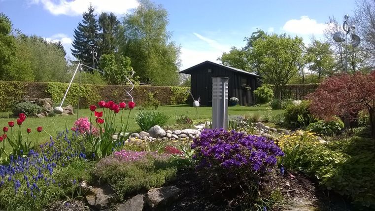 A beautiful garden with colorful flowers and a wooden house in the background. The green landscape is brightly lit by the sun.