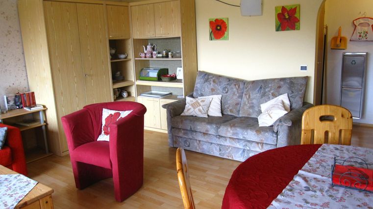 Ein gemütliches Wohnzimmer mit einem grauen Sofa, einem roten Sessel und einem Tisch. Die Wände sind hell und mit Bildern dekoriert.