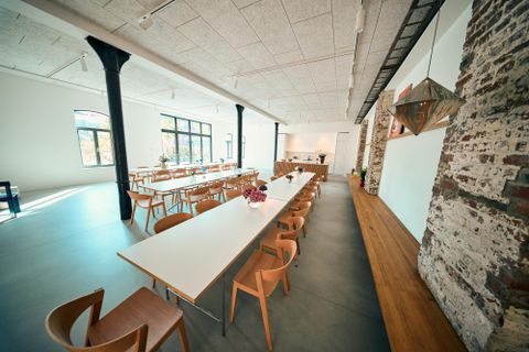 Ein großzügiger Raum mit langen Tischen und modernen Holzstühlen. Die Wände sind partly aus Ziegel und das Licht strömt durch große Fenster.