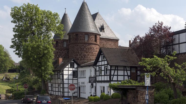 Ein historisches Gebäude mit Türmen und Fachwerkarchitektur. Umgeben von Bäumen und einem klaren Himmel.