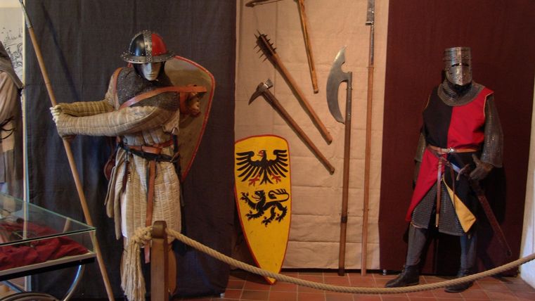 Een middeleeuws museum met twee figuren in wapenrusting. Op de achtergrond zijn verschillende wapens en een schild te zien.