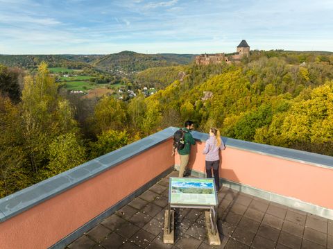 A view from a viewpoint with two people looking at the surrounding landscape. In the background, a castle ruin can be seen on a hill, surrounded by colorful trees.