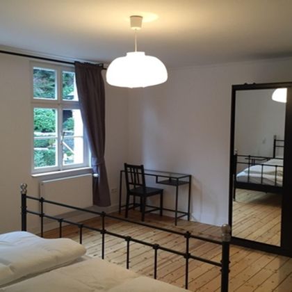 A simple bedroom with a double bed, a desk, and a large mirror. Bright walls and wooden flooring create a warm atmosphere.