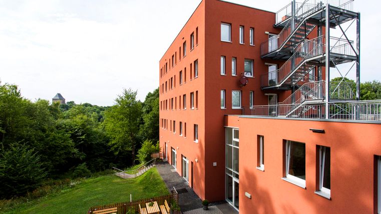 A modern red building with an outdoor exit and a fire escape. In the background, there are trees and a hilly landscape.