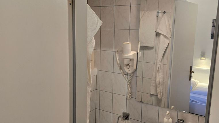 A small bathroom with a toilet, sink, and mirror. White tiles and towels are visible.