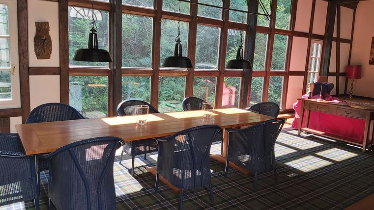 A bright, inviting room with a large table and chairs. Large windows let in plenty of daylight and offer a view of the greenery.