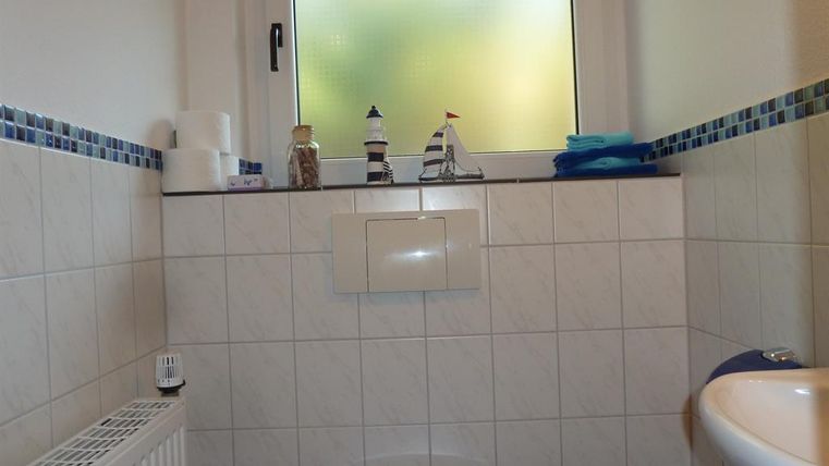 A simple bathroom with white tiles and a window with frosted glass. On the shelf are toilet paper, towels, and a decorative figure of a ship.