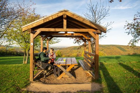 A cozy wooden gazebo on a meadow with a beautiful view. Two people are sitting at a table and having a conversation in nature.