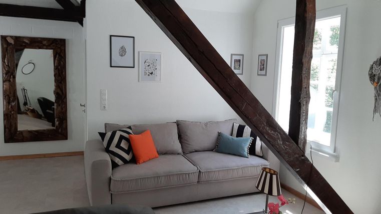 Een gezellige woonkamer met een grijze bank en kleurrijke kussens. De muren zijn deels versierd met houten lijsten, en er zijn grote ramen die veel licht binnenlaten.