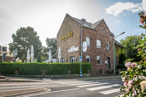 A cozy restaurant building at a street corner. Surrounded by green hedges and umbrellas, guests are seated outdoors.