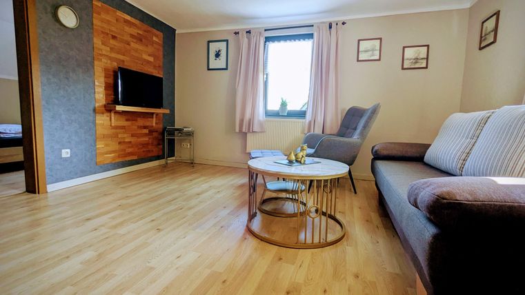 A cozy living room with wooden flooring and modern furniture. There is a sofa, a table, and a television on the wall.