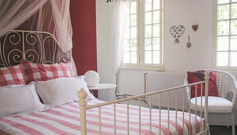 Ein gemütliches Schlafzimmer mit einem Bett mit rot-weißem Karomuster und weißen Kissen. Es gibt einen Stuhl und große Fenster, die viel Tageslicht hereinlassen.