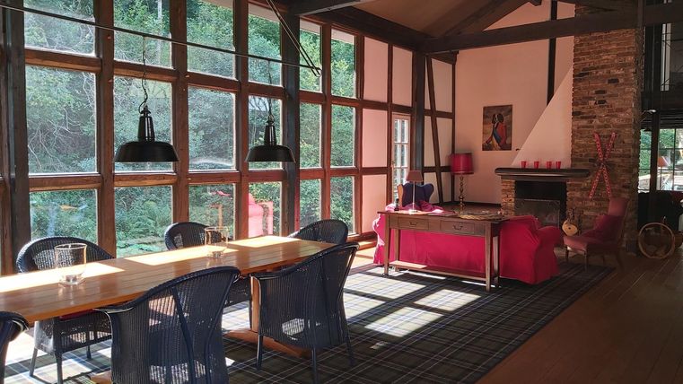A bright living room with large windows and plenty of daylight. There is a dining table, chairs, and a cozy area with a red sofa.