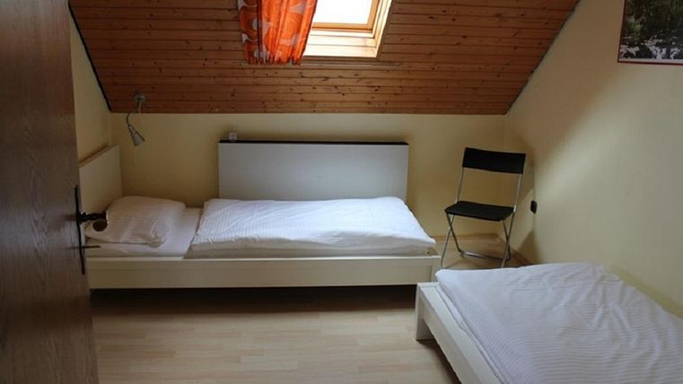 A simple room with two beds and a chair. It has a wooden ceiling and a window with an orange curtain.