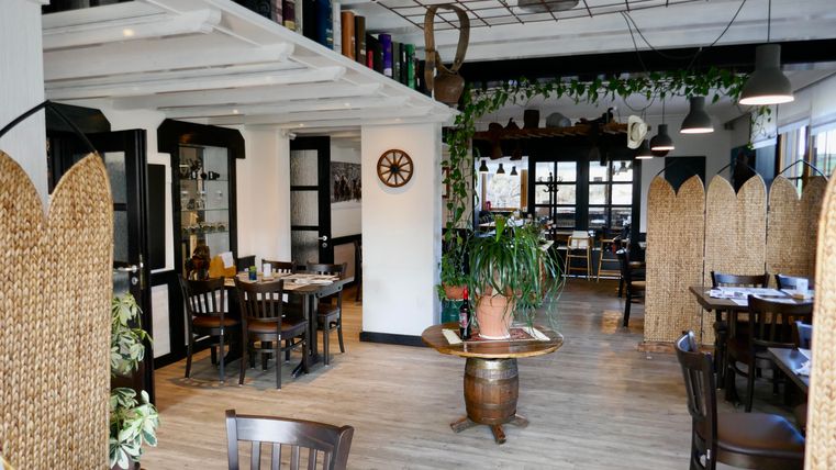 A cozy restaurant with wooden furniture and plants. The room is bright and inviting, with a warm atmosphere.