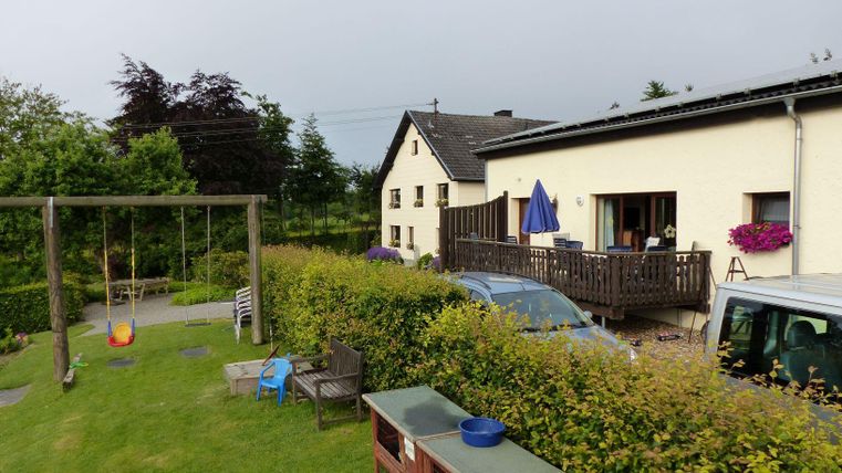 A quiet garden with a swing and a wooden deck. In the background stands a two-story house with blooming plants.