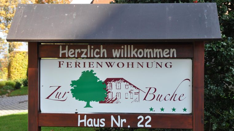 A welcome sign for a holiday apartment named "Zur Buche". It displays house number 22 and is surrounded by green plants.