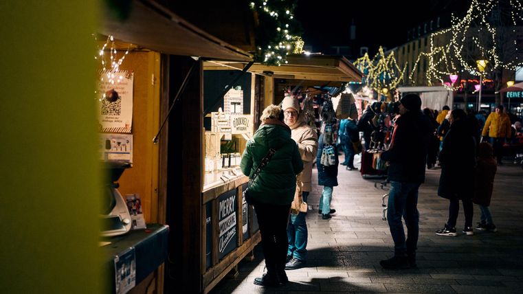 Ein belebter Weihnachtsmarkt bei Nacht mit festlicher Beleuchtung. Menschen stöbern an Holzständen mit Waren.