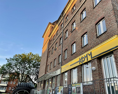 Ein charakteristisches Backsteingebäude mit gelber Beschilderung. Im Vordergrund sind Bäume und ein Eingang zu sehen.