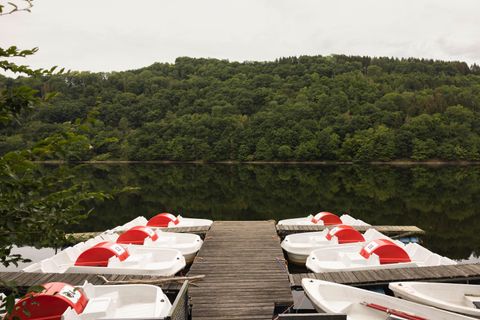 A tranquil lake with a dock equipped with pedal boats. In the background, green hills and a cloudy sky can be seen.