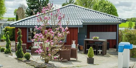 Een gezellig huis met een modern dak en een bloeiende boom op de voorgrond. De buitenruimte is ingericht met tuinmeubels en kleine planten.