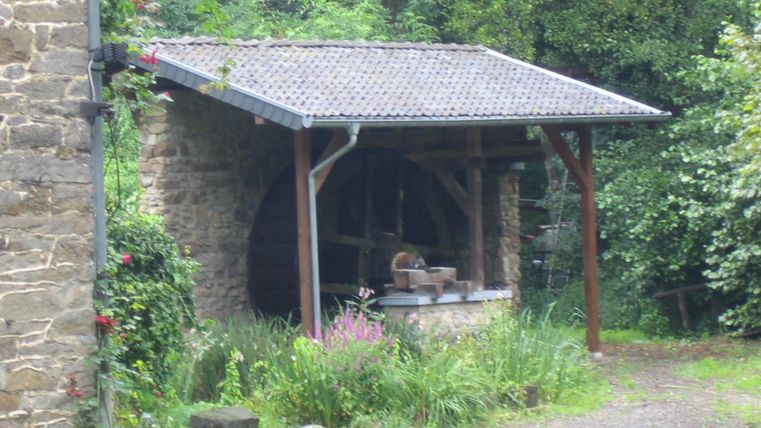Ein altes Mühlenhaus mit einem Wasserrad und grünem Gestrüpp. Die Umgebung ist ruhig und von Bäumen umgeben.