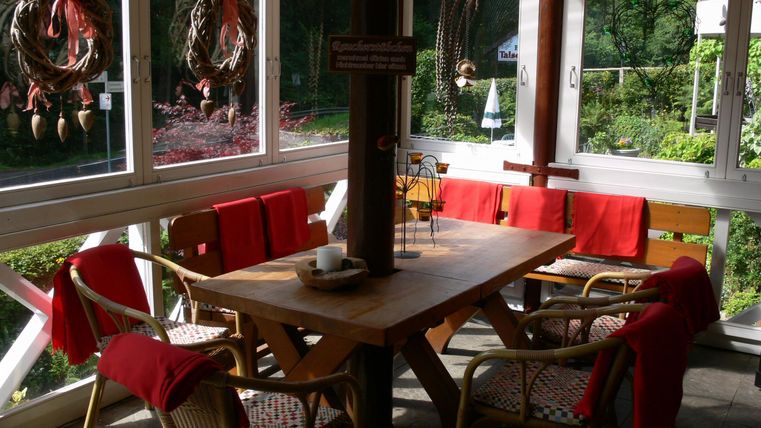 A cozy dining area with a wooden table and chairs, decorated with red blankets. Large windows offer a view of the green surroundings.