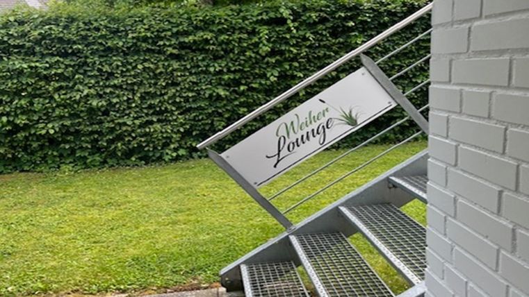 Een buitenaanzicht met een metalen trap en een bord dat wijst op de "Welker Lounge". Op de achtergrond zijn groene struiken en een verzorgd gazon te zien.