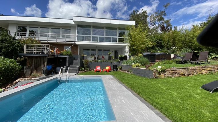Een modern huis met grote ramen en een verzorgde tuin. Op de voorgrond bevindt zich een zwembad, omgeven door gras en ligstoelen.