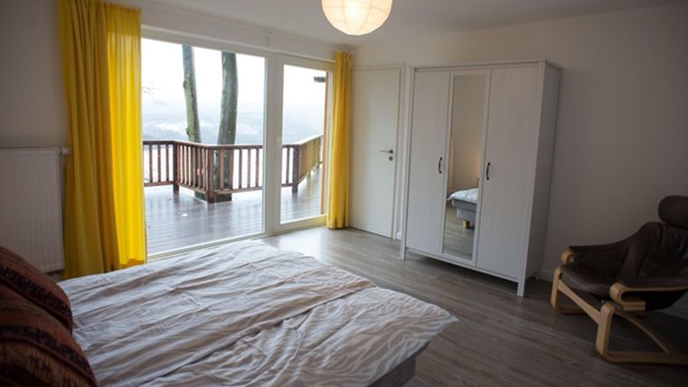 A bright bedroom with a large bed and a wardrobe. Large windows with yellow curtains offer a view of the terrace.