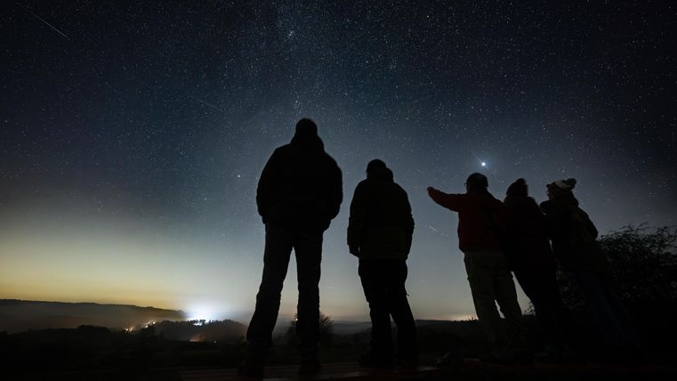 A group of people stands silhouetted against a clear night sky filled with stars. A gentle light glow is visible on the horizon.