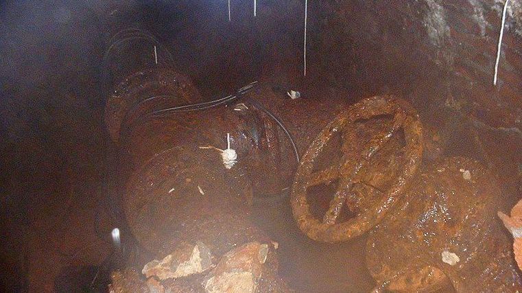 Eine verrostete Rohrleitung unter der Erde mit einer aufliegenden Ventilsteuerung. Der Bereich wirkt feucht und ist mit zerfallenen Ziegeln umgeben.