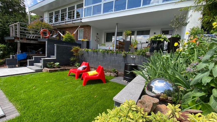 Een moderne woning met grote ramen en een verzorgde tuin. Op de voorgrond staan rode stoelen en verschillende planten die een uitnodigende sfeer creëren.